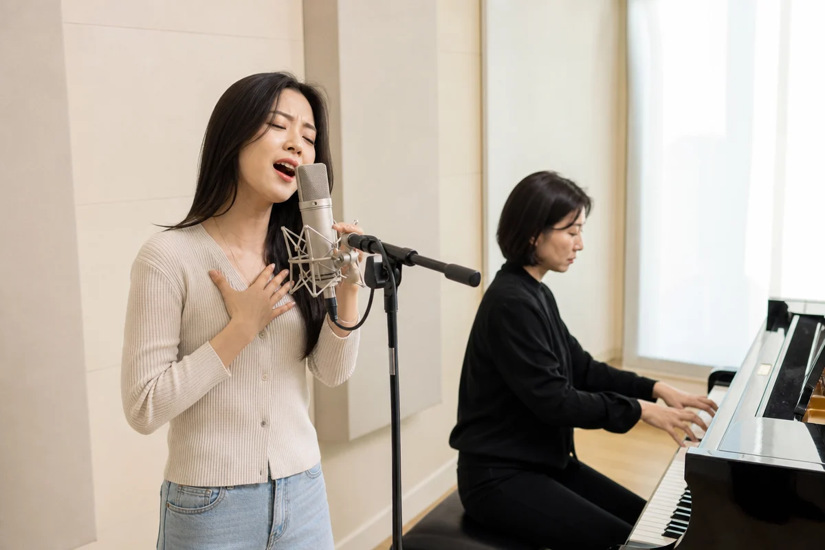 피아노 반주에 맞춰 노래 연습하는 수강생과 강사의 보컬레슨 비용 상담 장면